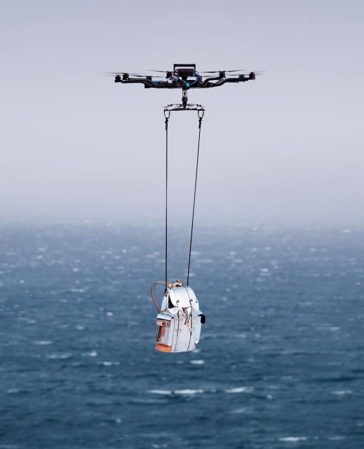 Drone carries a white backpack by ropes over a dark blue ocean under an overcast sky, aerial technology with nature backdrop.