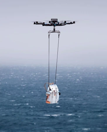 Drone carries a white backpack by ropes over a dark blue ocean under an overcast sky, aerial technology with nature backdrop.