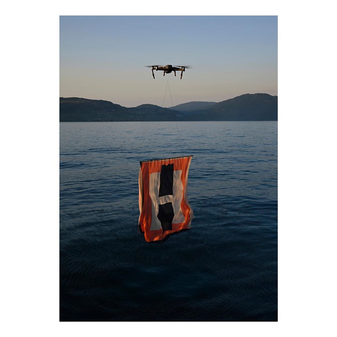 Drone flying over water carries an orange and black banner in a scenic outdoor setting with mountains in the distance under a serene sky.