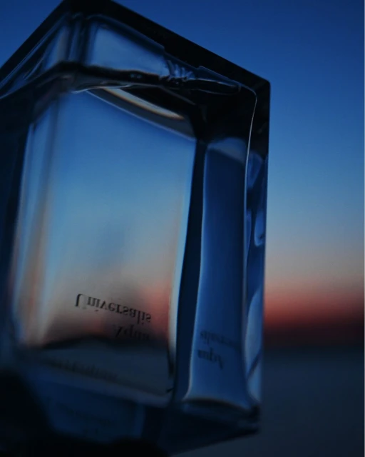 A close-up studio shot captures a rectangular blue perfume bottle against a gradient sky backdrop with twilight hues.
