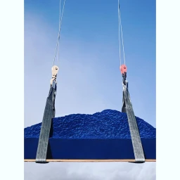 Blue textured object suspended in the air by heavy duty industrial straps and metal cables against a clear blue sky.