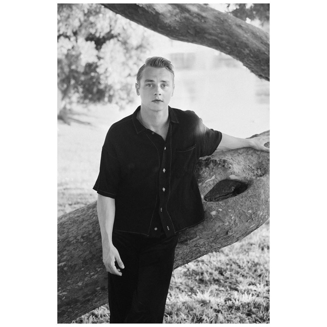 Monochrome portrait of a blond adult man in a dark shirt leaning against a fallen tree in a grassy outdoor setting.
