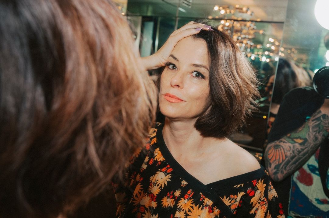 Woman with short brunette hair poses looking in a mirror, wearing a floral print wrap top in an indoor environment with warm lighting.