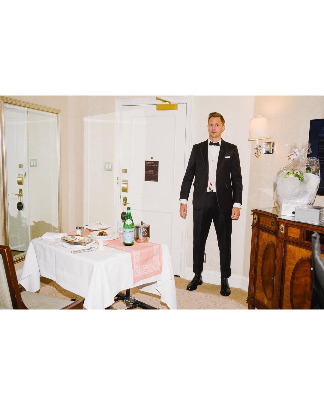 Adult man in tuxedo poses in hotel room near white-tabled meal service, sideboard with bouquet, mirrored doors, and neutral decor.