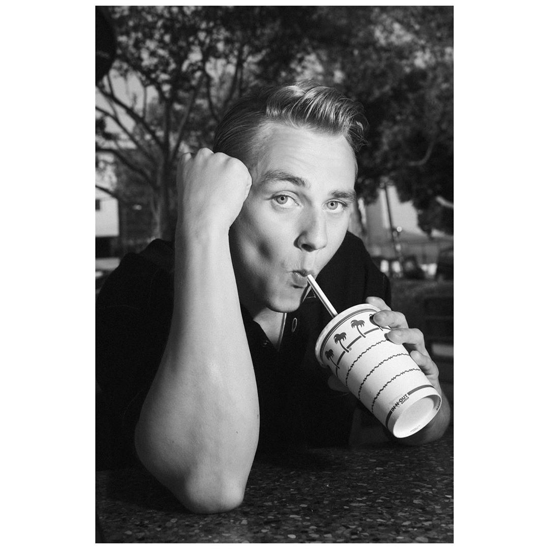Monochromatic portrait of a young adult male drinking from a paper cup with a straw, with trees and a building in the background.