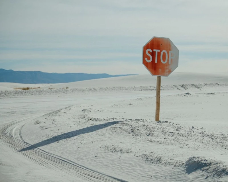 Stop sign stands alone in the white sand dunes, with tire tracks visible. Mountains are faintly visible in the distance under a blue sky.