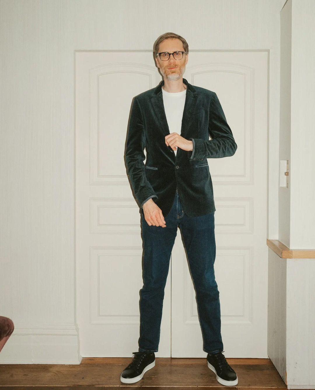Full-length portrait of an adult male wearing a velvet blazer, t-shirt, and jeans, standing indoors against a white wall and doorway.