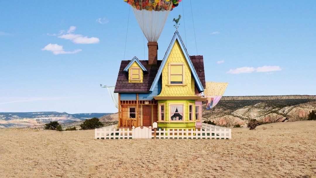 Whimsical cartoon house floats in a desert landscape beneath a canopy of colorful balloons against a clear blue sky. Day.