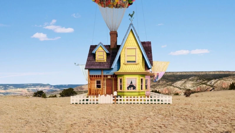 Whimsical cartoon house floats in a desert landscape beneath a canopy of colorful balloons against a clear blue sky. Day.