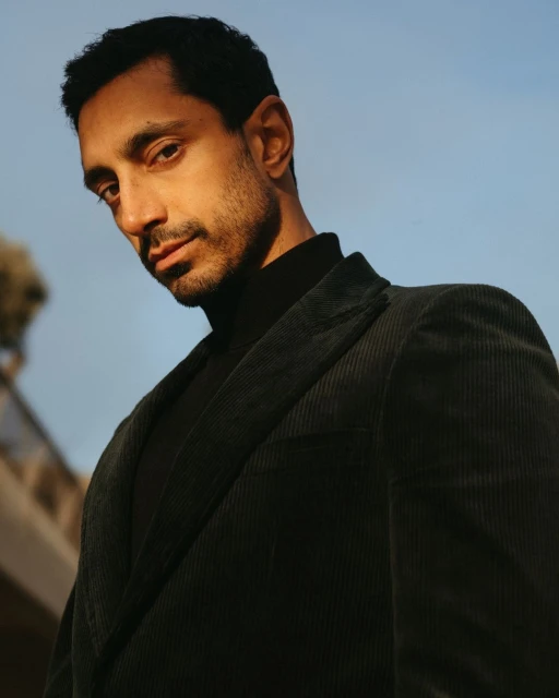 Portrait of a man in a dark corduroy blazer against a blue sky. He is posing at a low angle looking down at the camera.