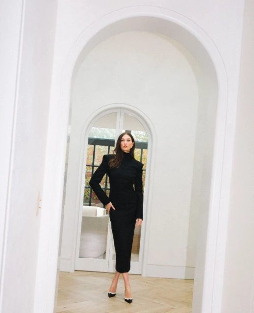 A poised woman in a black midi dress with structured shoulders stands in a bright white doorway. She completes her look with pointed heels.