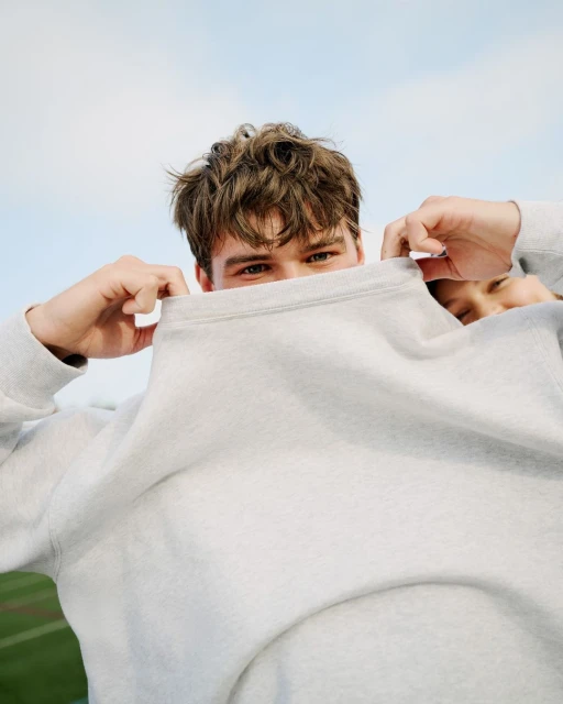 Close-up of a young adult covering their face with a gray sweatshirt against a bright sky, showcasing casual style and outdoor freshness.