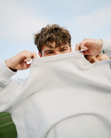 Close-up of a young adult covering their face with a gray sweatshirt against a bright sky, showcasing casual style and outdoor freshness.