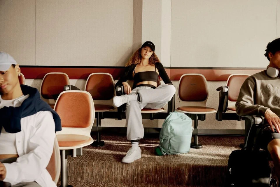 Three people sit in a waiting room, one woman in the center wearing casual clothing, a backpack at her feet. Calm indoor scene.