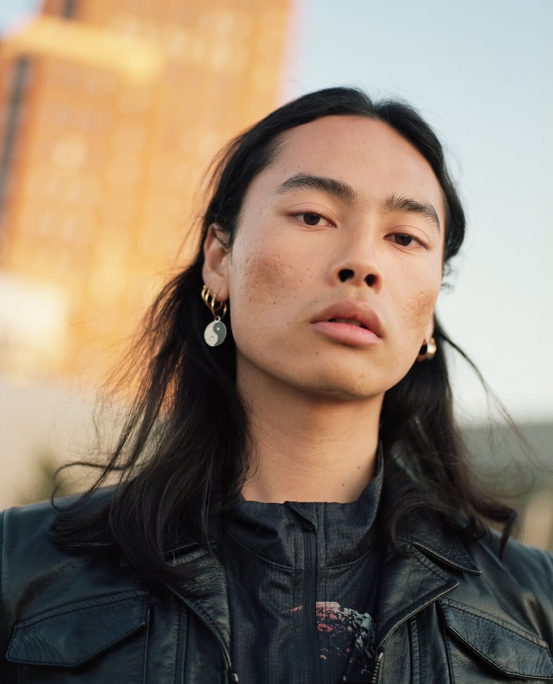 Portrait of an androgynous person with long dark hair wearing a black leather jacket, against a backdrop of tall buildings.