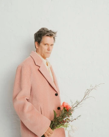 Portrait of actor Kevin Bacon wearing a pink corduroy coat and holding a small bouquet of flowers against a white wall.