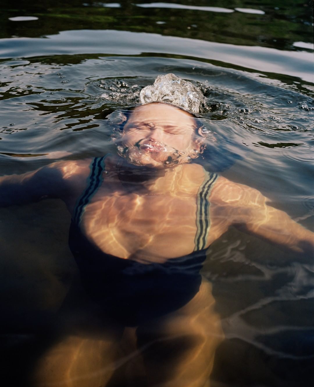 Woman in a one-piece swimsuit swimming in a lake with a tree-lined shore in the background, sun reflecting on the water's surface.