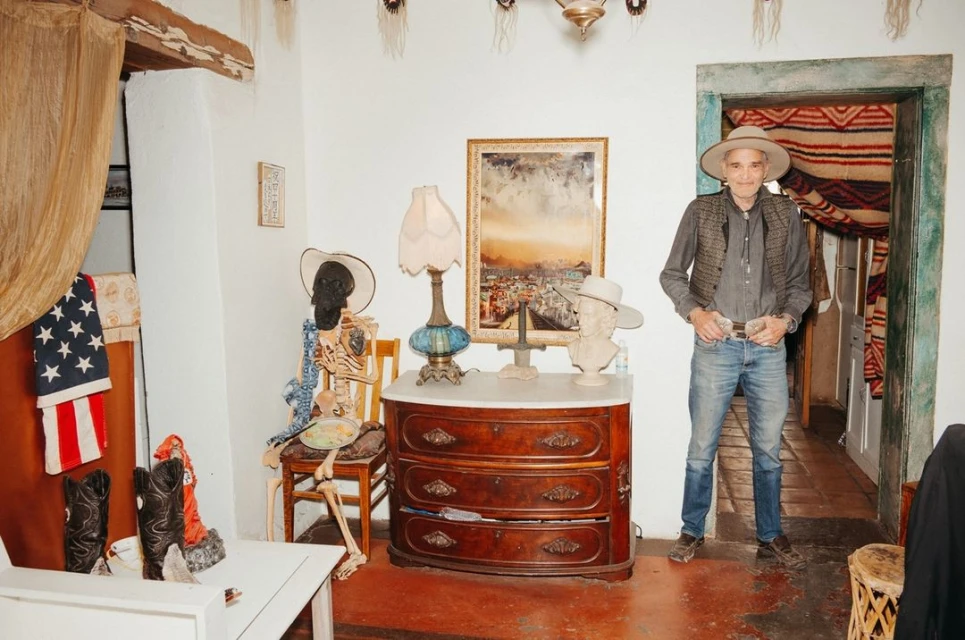 Full indoor shot of an adult male in a cowboy hat and vest, standing near a dresser with cowboy boots and a skeleton on a chair.