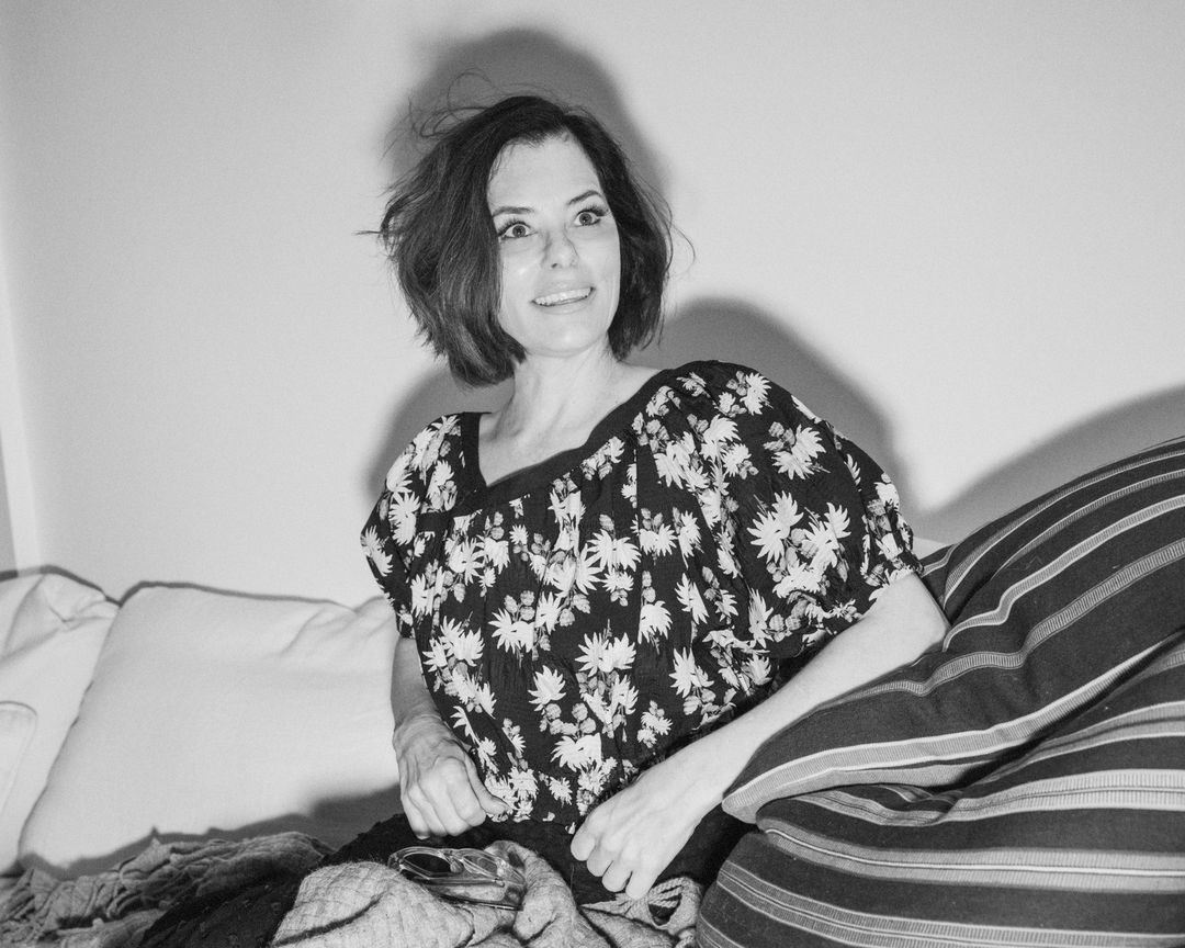 Portrait of an adult woman with dark hair, wearing a black and white floral top, smiling and sitting with pillows against a wall.