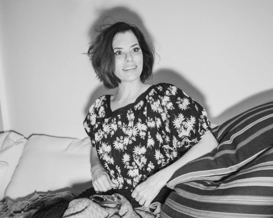 Portrait of an adult woman with dark hair, wearing a black and white floral top, smiling and sitting with pillows against a wall.
