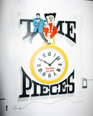 Vintage 'Time Pieces Est. 1979' sign with cartoon figures and a clock, mounted on a white wall, showcasing retro design and functionality.