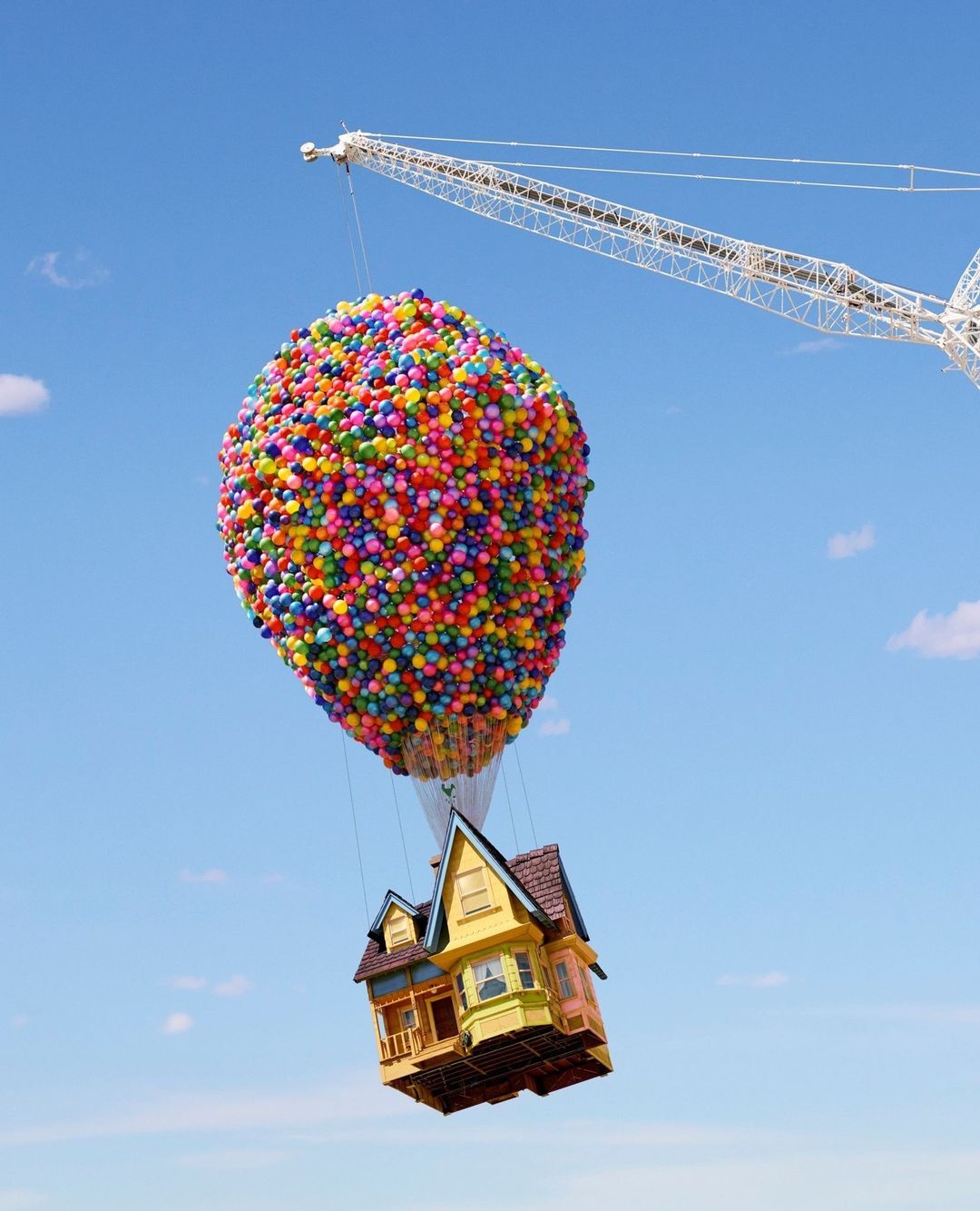 House suspended by colorful balloons against a blue sky, attached to crane. Dreamlike scene with childhood nostalgia.