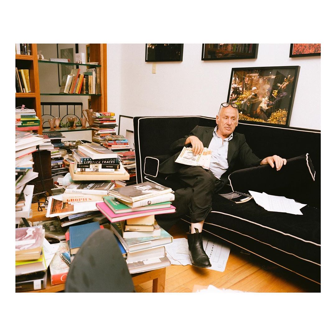 Mature man sitting on a black couch in a room filled with books, wearing a dark suit, holding a magazine in his hand.