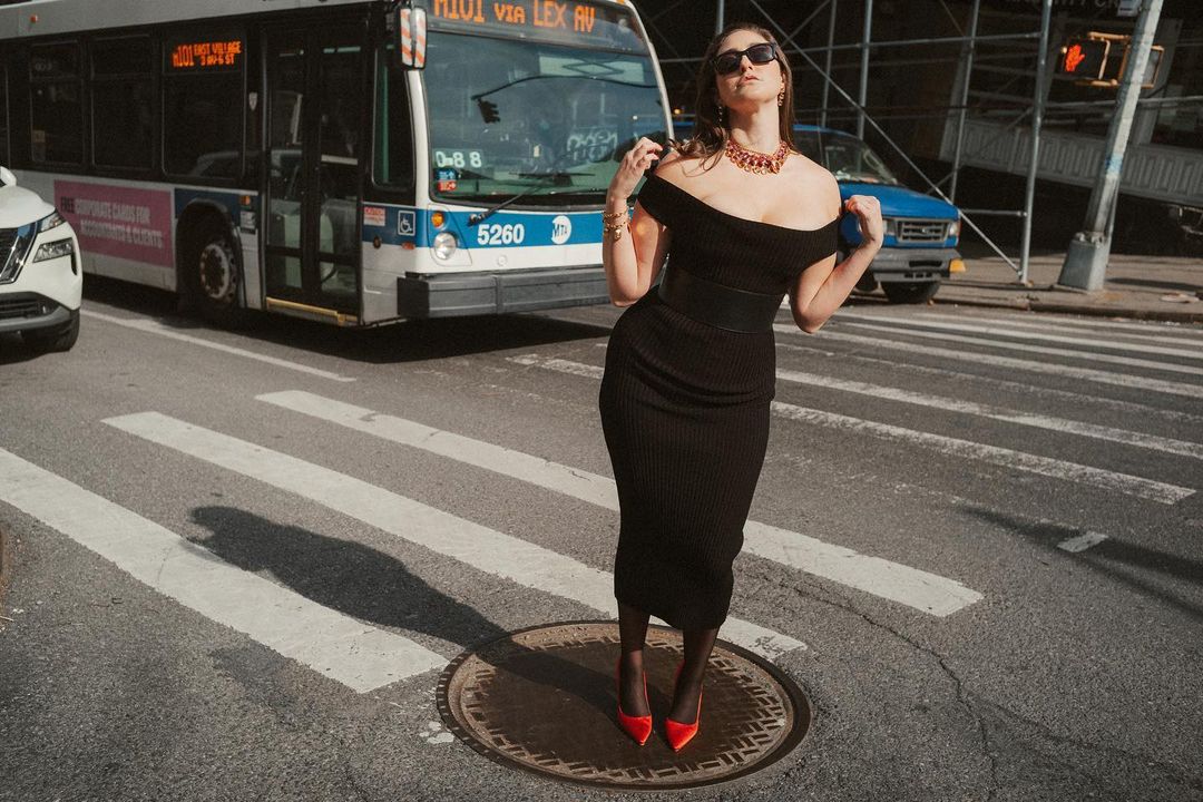 Woman in black dress standing on a crosswalk with red heels and statement necklace, posing in front of a bus in a city street.