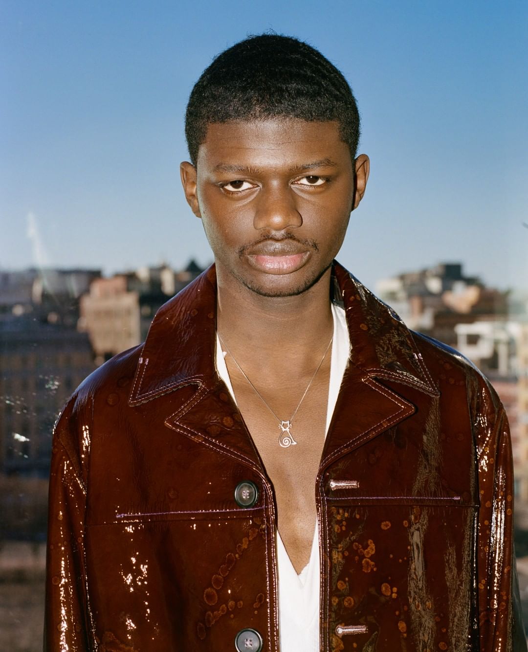 Portrait of an adult male with short dark hair, wearing a shiny brown jacket and necklace, posing against a city skyline and blue sky.