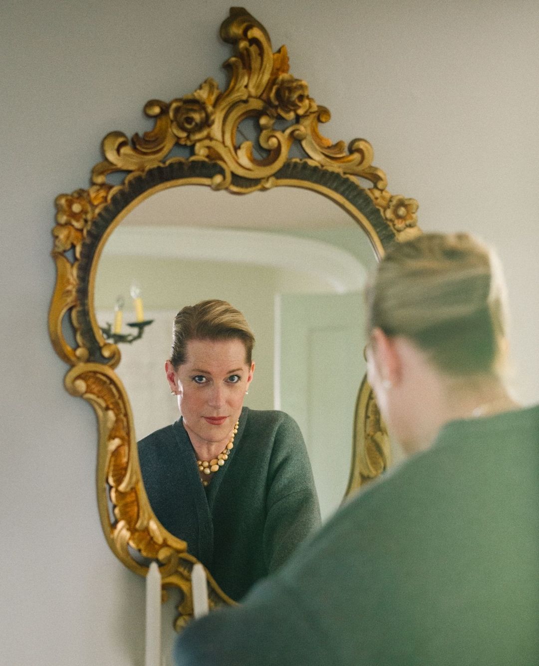 Woman reflected in an ornate gold mirror, wearing a dark green sweater, pearl necklace, and earrings; indoor environment with a white wall.