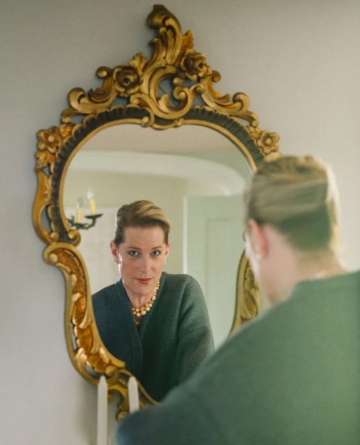 Woman reflected in an ornate gold mirror, wearing a dark green sweater, pearl necklace, and earrings; indoor environment with a white wall.