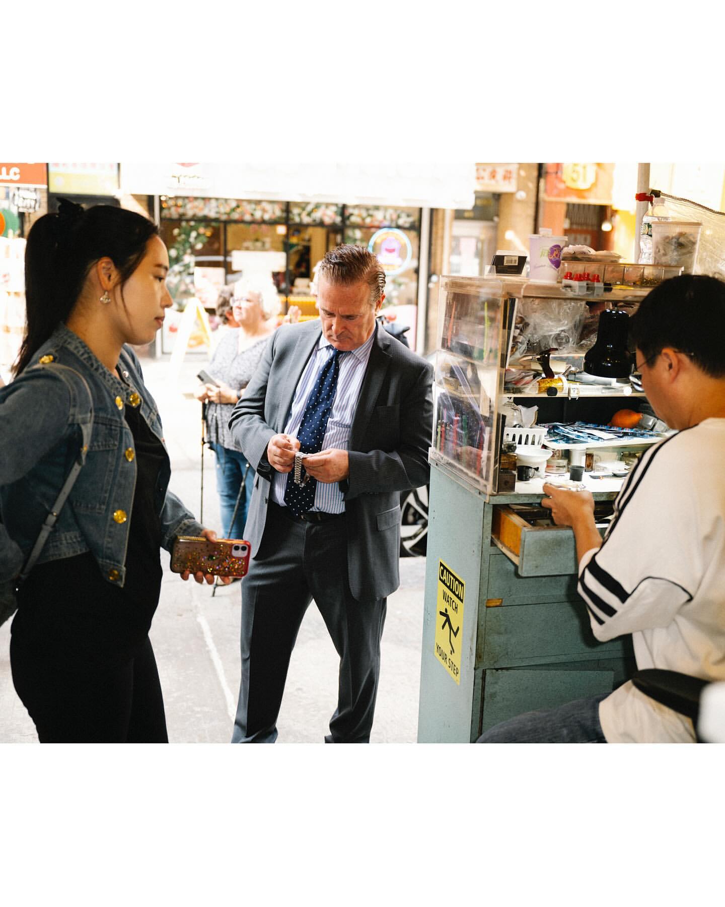 Outdoor scene with three people: a woman in a denim jacket, a man in a suit paying at a street vendor stall, and another man behind the stall.