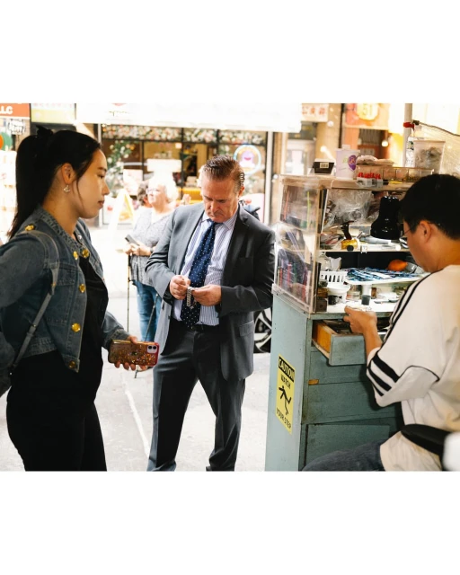 Outdoor scene with three people: a woman in a denim jacket, a man in a suit paying at a street vendor stall, and another man behind the stall.