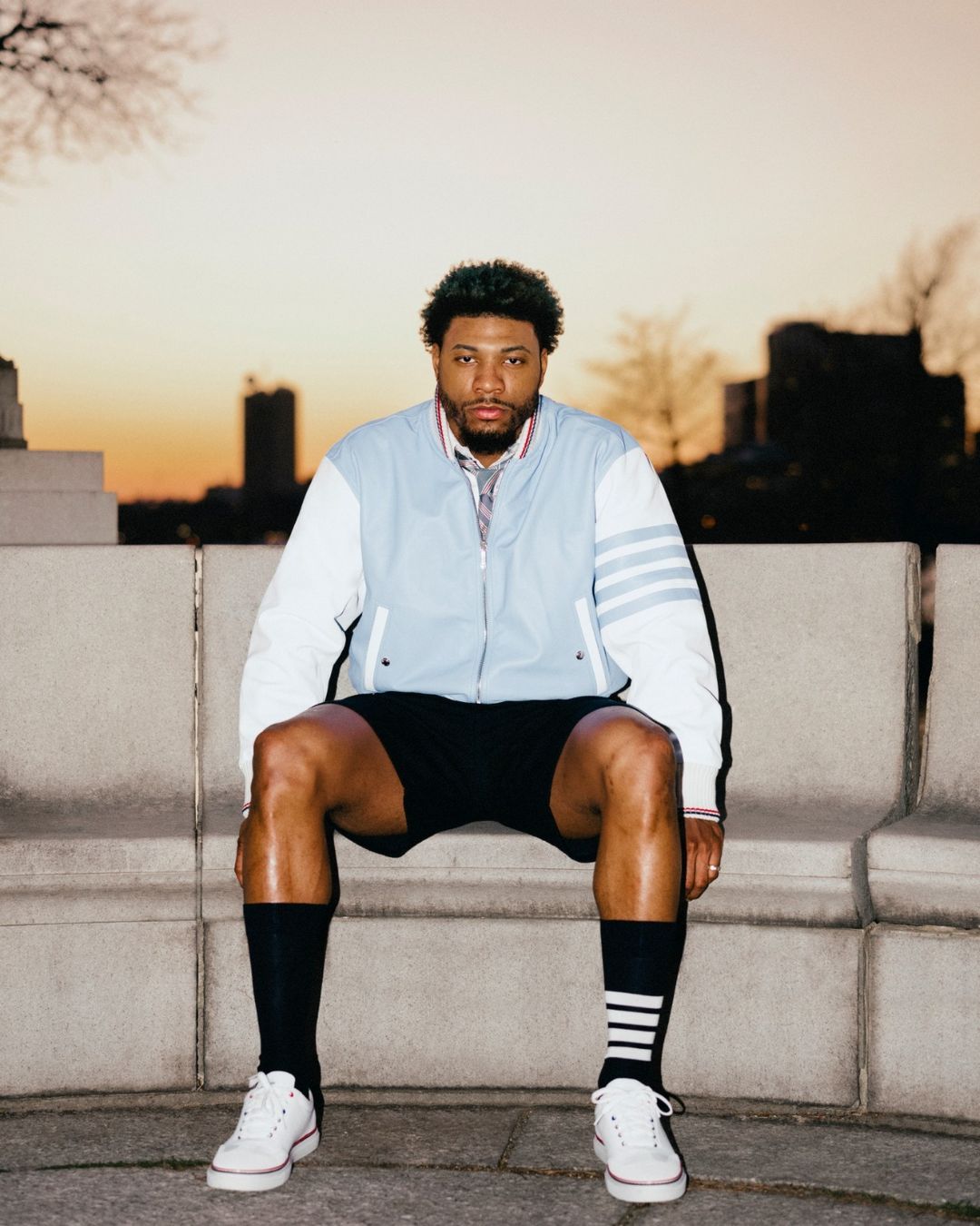 Man seated on concrete ledge wearing light blue varsity jacket, black shorts, and white sneakers against sunset sky.