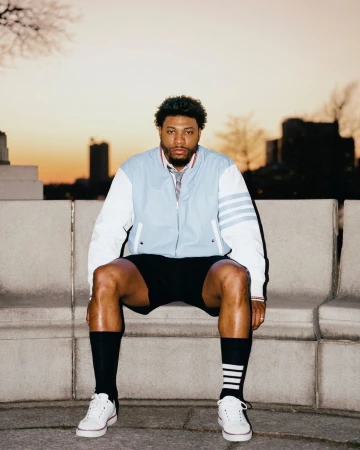 Man seated on concrete ledge wearing light blue varsity jacket, black shorts, and white sneakers against sunset sky.