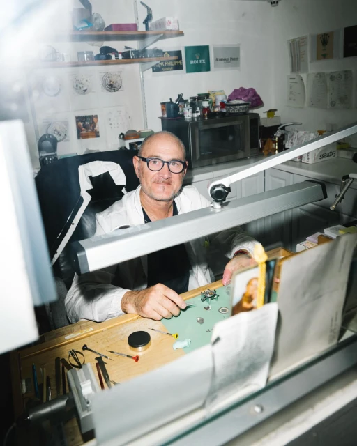 Watchmaker at workbench repairing watches with parts, tools, and magnifying lamp in a bright workspace, wearing glasses and a lab coat.