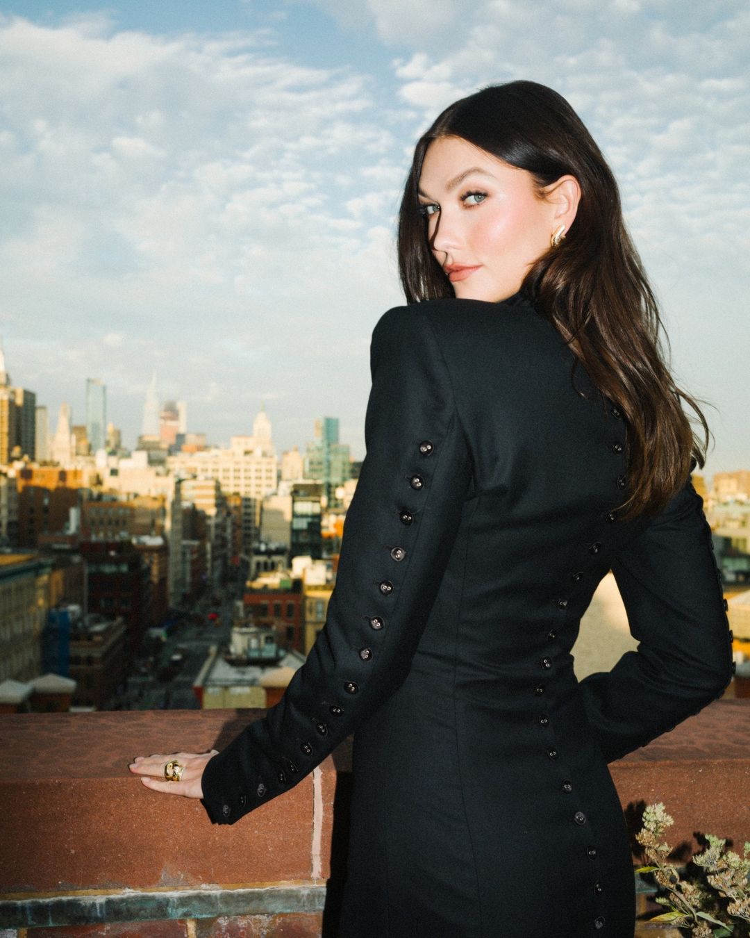 Model Karlie Kloss in a black button-detail dress looking back on a rooftop overlooking a city skyline by photographer Ryan Lowry
