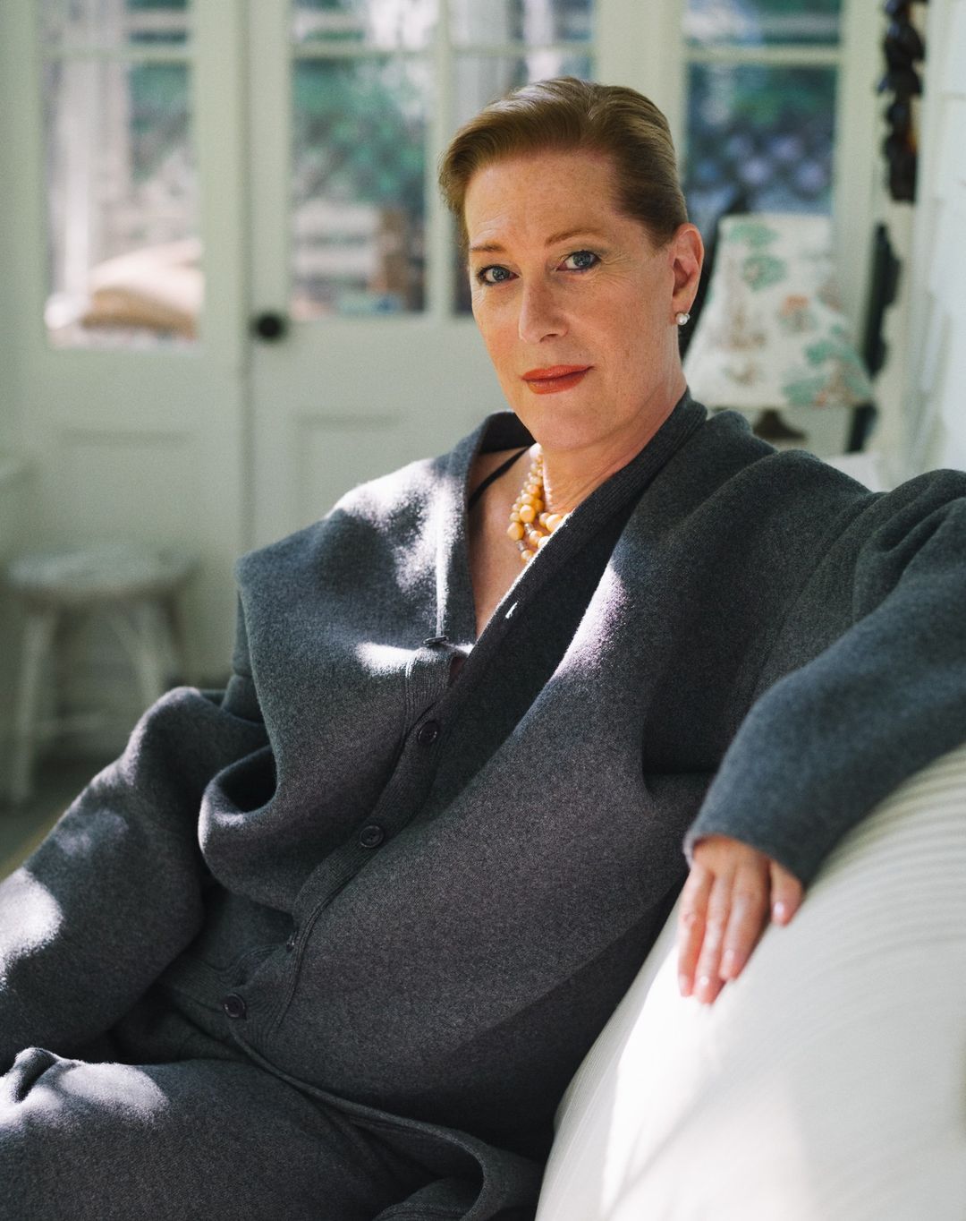 Portrait of a mature woman with short hair, wearing a grey outfit and pearl necklace, sitting on a white sofa in a light indoor setting.
