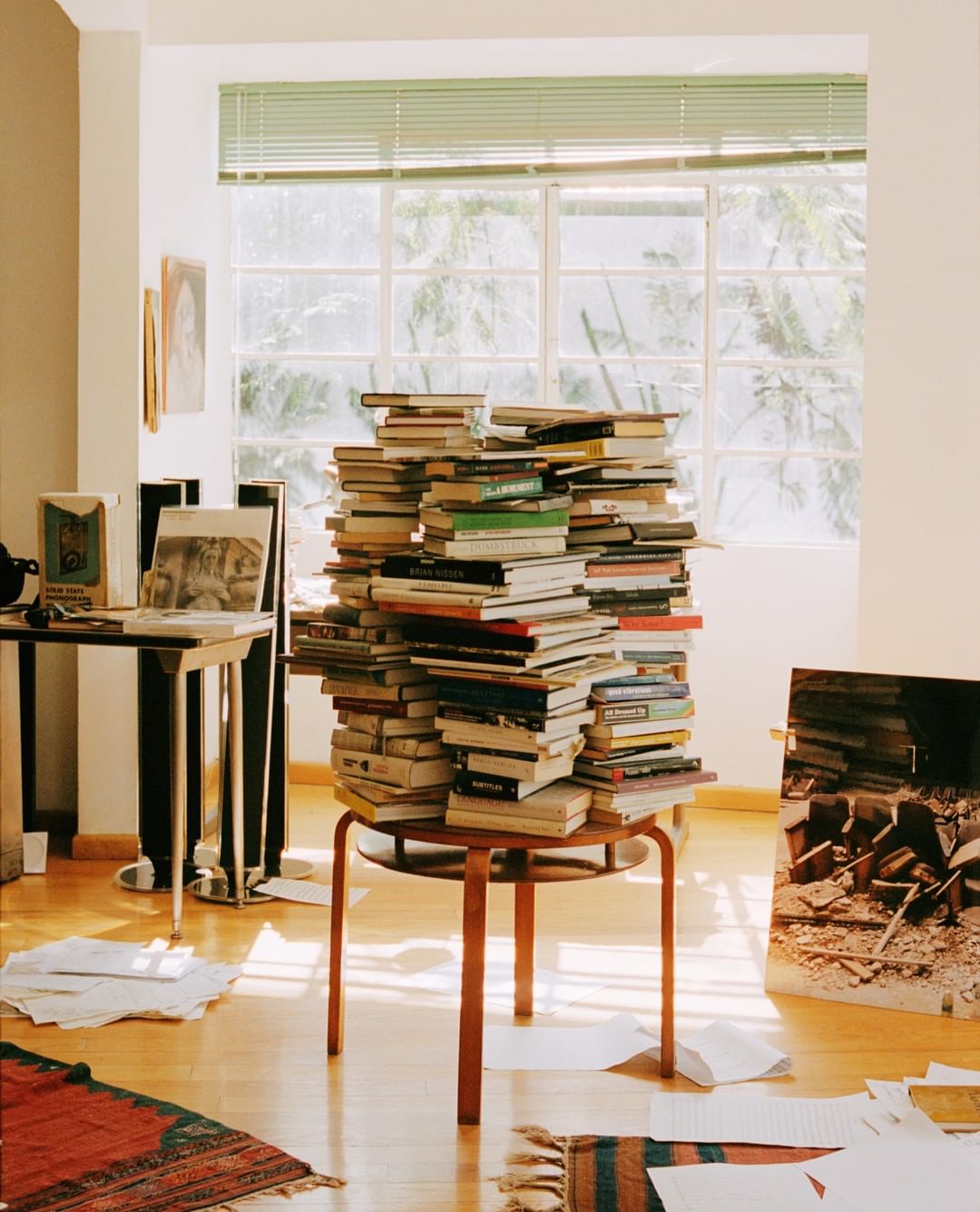 A tall stack of books sits on a wooden stool in a sunlit room with a table, framed picture, rug, and scattered papers on a wood floor.