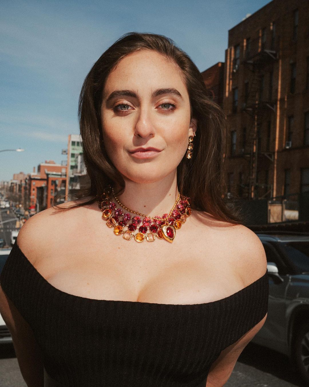 A young adult woman with long brown hair poses outdoors wearing a black off-the-shoulder dress and a gemstone necklace, with urban background.