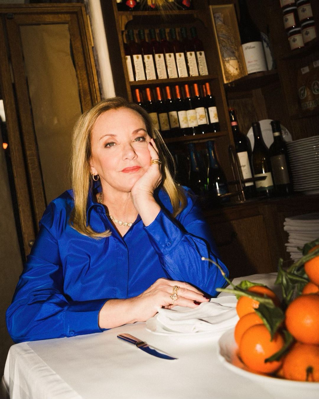 Portrait of an adult woman with blonde hair wearing a royal blue shirt, sitting at a table with oranges, in a restaurant setting.