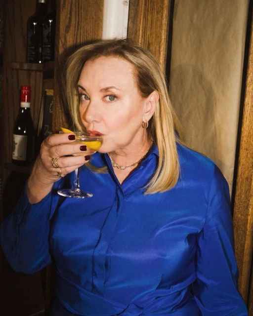 Woman drinks cocktail in front of wine rack. She wears a bright blue blouse with gold jewelry in an indoor setting with warm lighting.