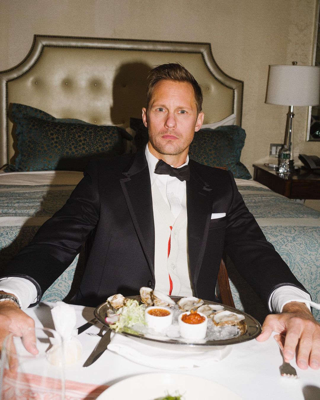 Adult male in a black tuxedo sitting at a table with a plate of oysters in front of him in an indoor setting.