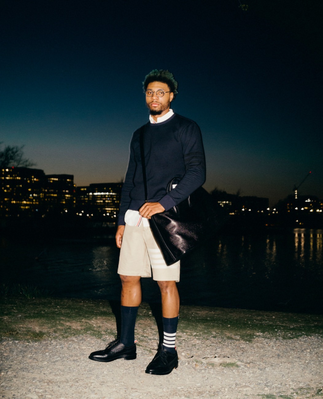 An adult male stands outdoors at dusk near a river wearing a navy sweater, khaki shorts, dark socks with stripes, and black dress shoes.