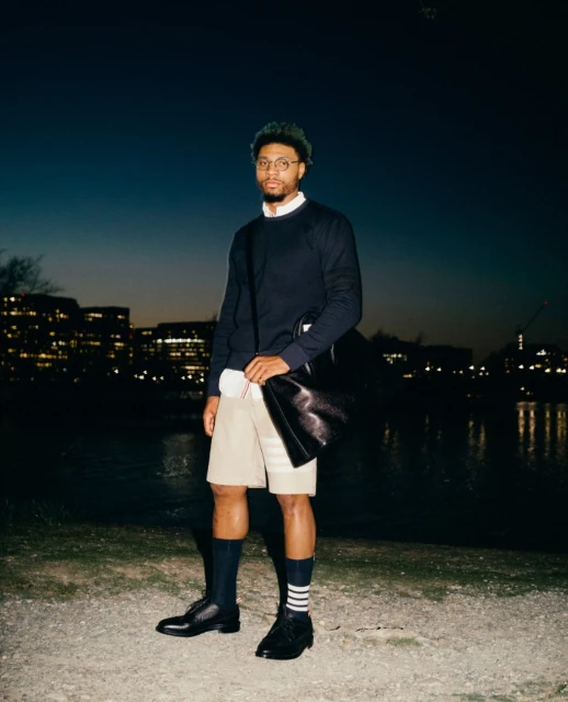 An adult male stands outdoors at dusk near a river wearing a navy sweater, khaki shorts, dark socks with stripes, and black dress shoes.