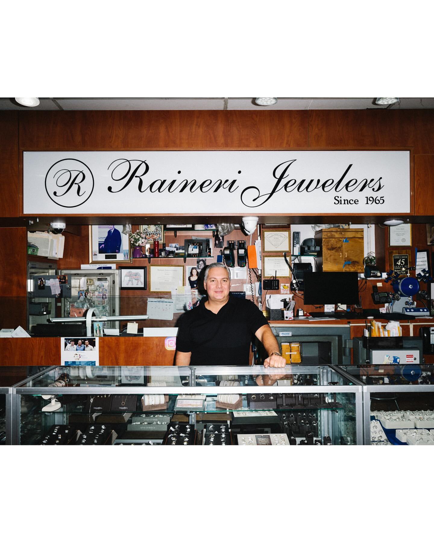 Man stands behind a jewelry store display case filled with rings, necklaces, and bracelets under a Rainier Jewelers sign.