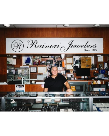 Man stands behind a jewelry store display case filled with rings, necklaces, and bracelets under a Rainier Jewelers sign.
