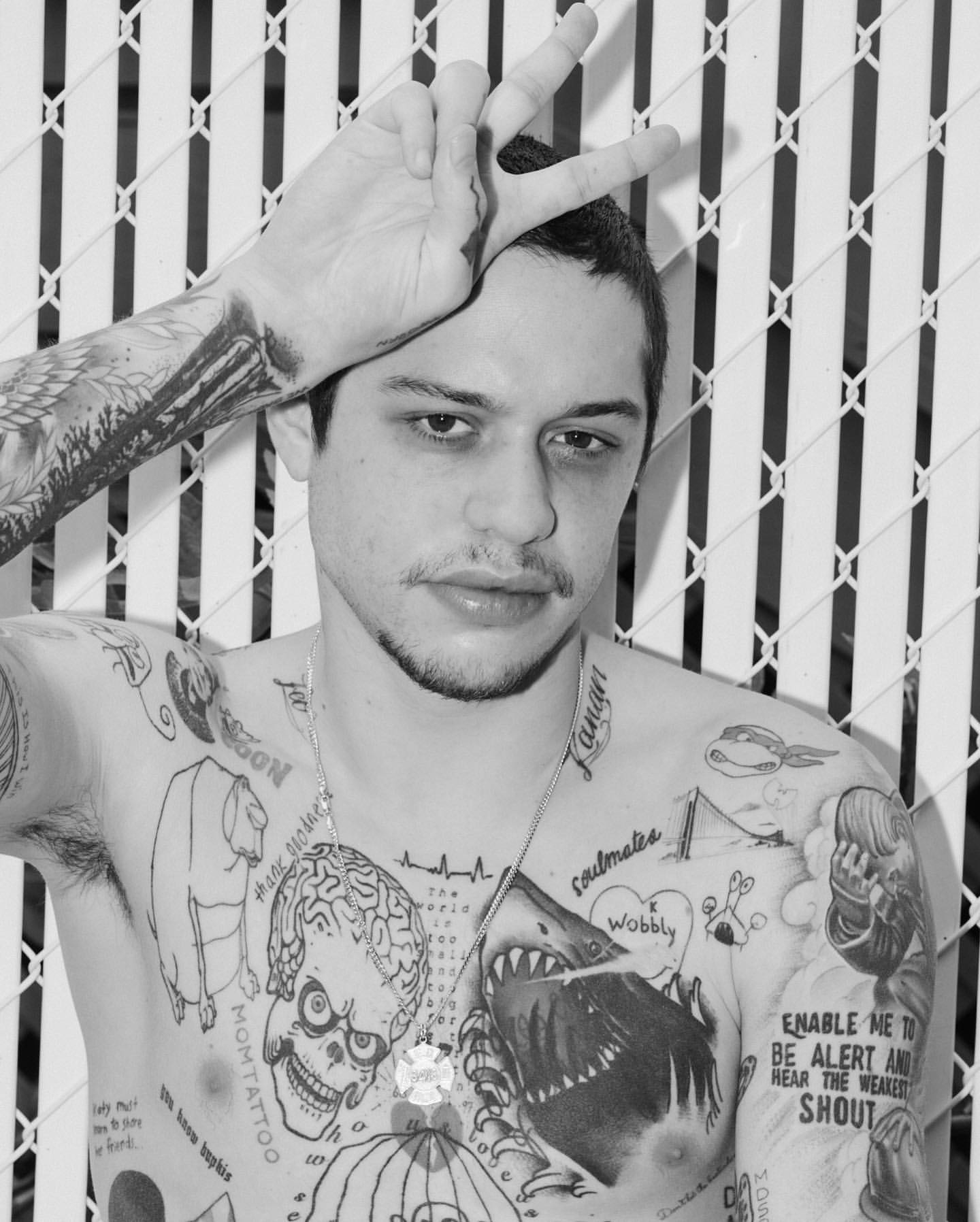 Monochrome portrait of a shirtless adult male with tattoos making a peace sign in front of a fence. Shallow depth of field.