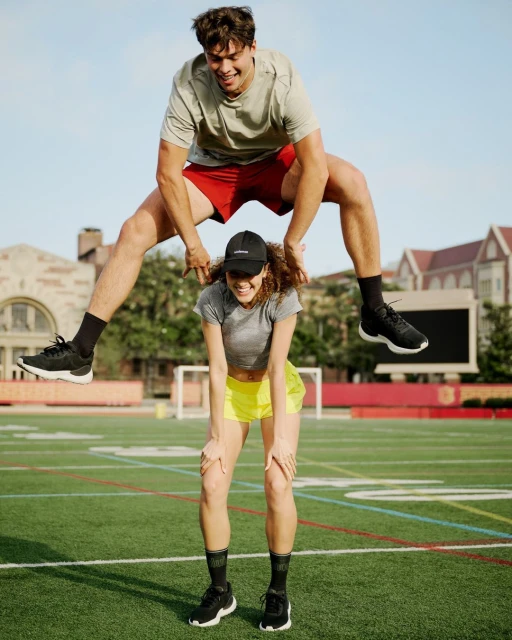 Man jumps over a woman's shoulders on a green field, both smiling. The man wears a tee and red shorts, the woman a gray tee and yellow shorts.