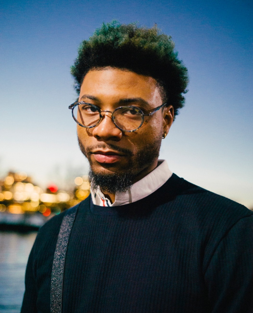 Portrait of a young Black man with round glasses and dark curly hair with blue tips, wearing a sweater with a blurry city background.
